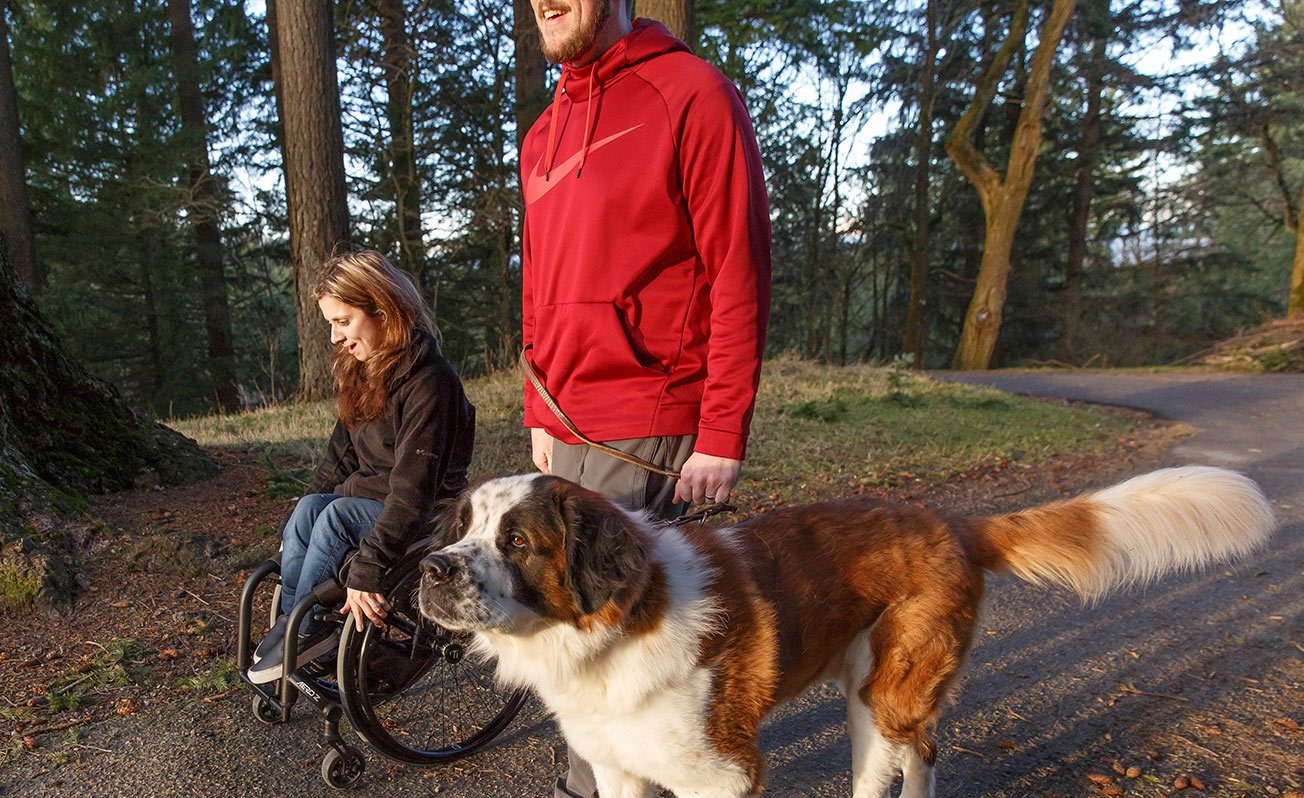 A woman in a wheelchair and a man with a large dog enjoy a trail in the sun